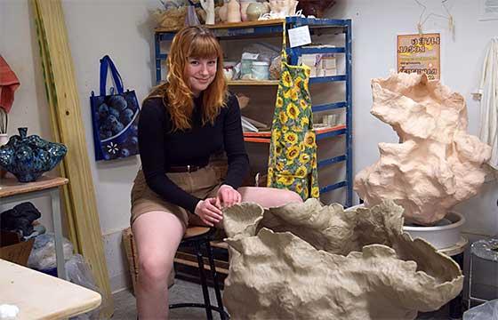 萨拉坐在工作室的凳子上，身后是正在制作的雕塑